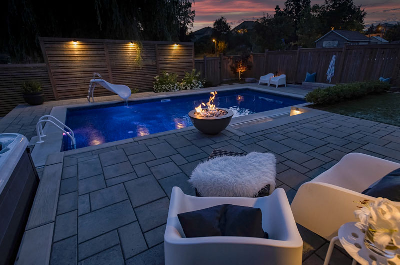 Nature-inspired, luminescent backyard boasting a large pool, hot tub, fire pit-lit patio. Minimalist, soft-focus blend of traditional-modern.